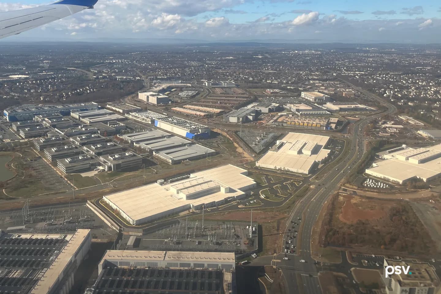 A daylight aerial photograph over Loudoun County, Virginia, looking across a dense field of low, flat-roofed data center buildings in pale grey and white, arranged in long parallel rows with rooftop cooling equipment, separated by wide access roads and parking aprons; two fenced electrical substations with lattice steel structures and busbars sit in the near foreground, a multi-lane divided highway runs diagonally through the frame at right past bare red-brown graded ground, and low suburban rooftops and tree cover stretch to a hazy ridgeline on the horizon. A daylight aerial photograph over Loudoun County, Virginia, looking across a dense field of low, flat-roofed data center buildings in pale grey and white, arranged in long parallel rows with rooftop cooling equipment, separated by wide access roads and parking aprons; two fenced electrical substations with lattice steel structures and busbars sit in the near foreground, a multi-lane divided highway runs diagonally through the frame at right past bare red-brown graded ground, and low suburban rooftops and tree cover stretch to a hazy ridgeline on the horizon.