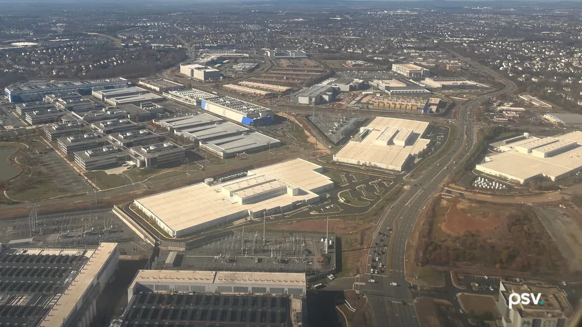 A daylight aerial photograph looking down over a Northern Virginia commercial corridor, with rows of long, low, windowless data center buildings with pale flat roofs and rooftop cooling units set among grey office blocks, a fenced electrical substation of steel lattice and transformers in the left foreground, a second substation beside a large pale warehouse at centre, a divided multi-lane highway curving through the frame at right with light traffic, bare winter trees and patches of graded red-brown earth between the lots, and suburban rooftops receding to a hazy ridgeline on the horizon.