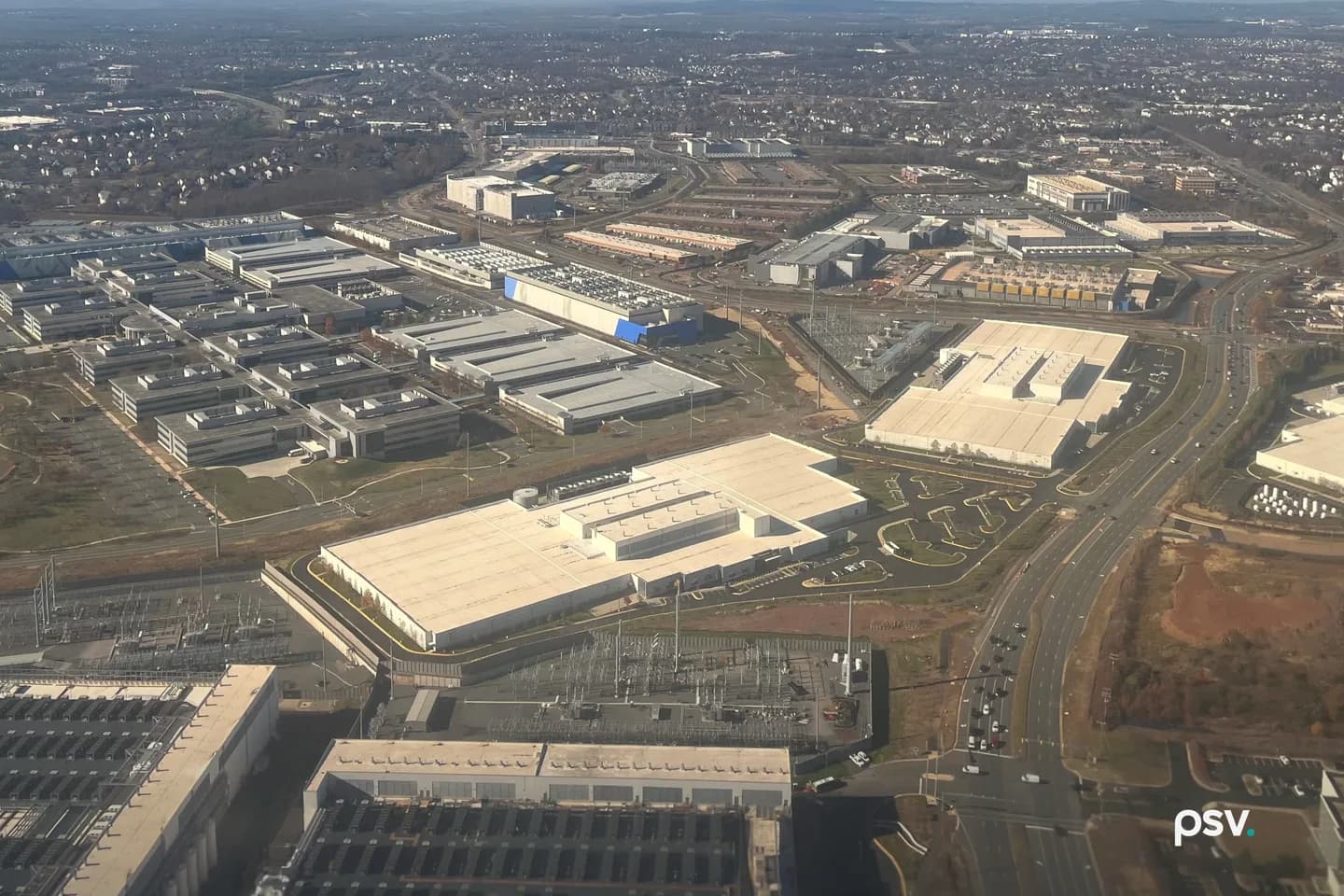 A daylight aerial photograph looking down over a Northern Virginia commercial corridor, with rows of long, low, windowless data center buildings with pale flat roofs and rooftop cooling units set among grey office blocks, a fenced electrical substation of steel lattice and transformers in the left foreground, a second substation beside a large pale warehouse at centre, a divided multi-lane highway curving through the frame at right with light traffic, bare winter trees and patches of graded red-brown earth between the lots, and suburban rooftops receding to a hazy ridgeline on the horizon. A daylight aerial photograph looking down over a Northern Virginia commercial corridor, with rows of long, low, windowless data center buildings with pale flat roofs and rooftop cooling units set among grey office blocks, a fenced electrical substation of steel lattice and transformers in the left foreground, a second substation beside a large pale warehouse at centre, a divided multi-lane highway curving through the frame at right with light traffic, bare winter trees and patches of graded red-brown earth between the lots, and suburban rooftops receding to a hazy ridgeline on the horizon.