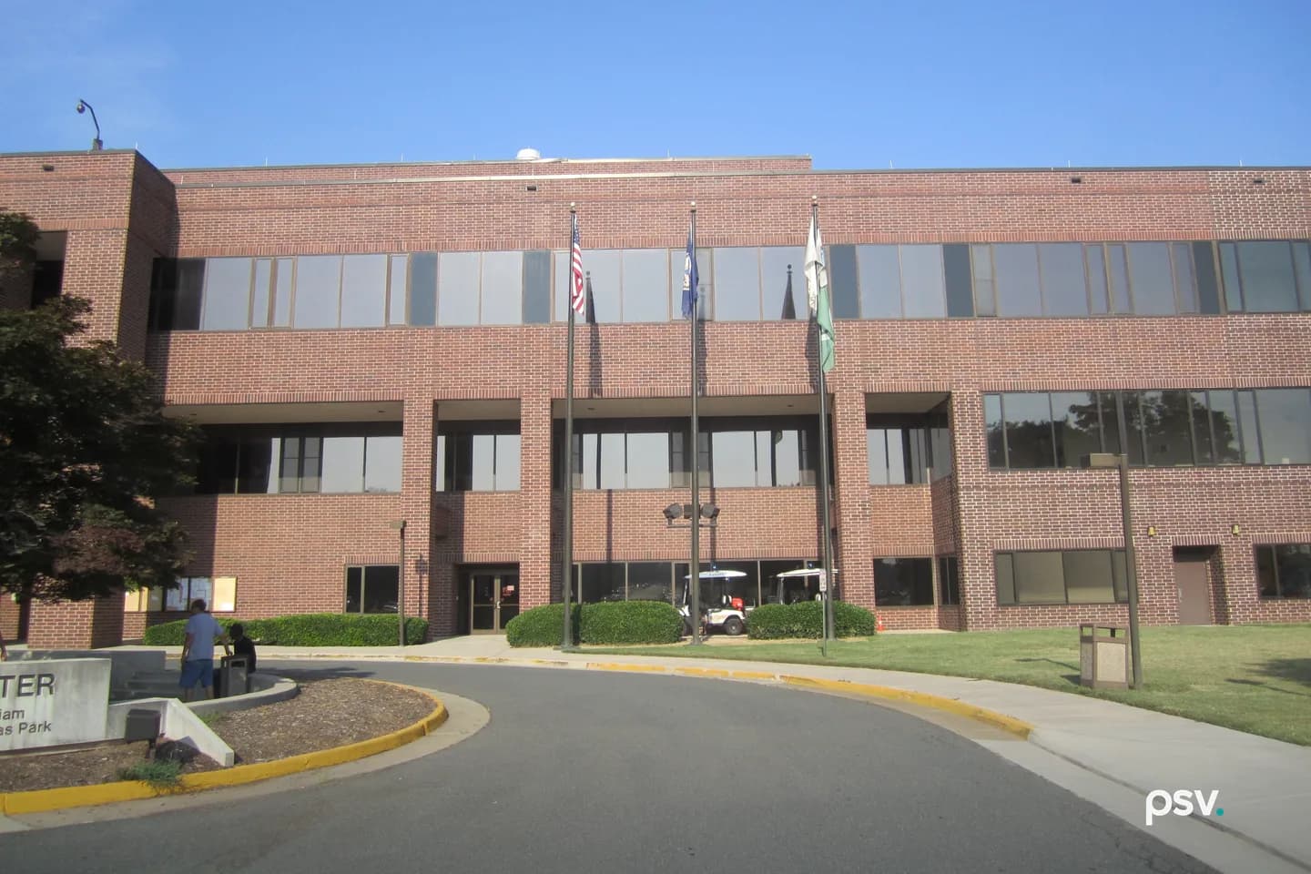 A daylight photograph of a two-storey red brick government building with continuous bands of tinted windows, a recessed entrance at the centre, three flagpoles flying the United States, Virginia and county flags on the lawn in front, clipped shrubs along the facade and a curving access drive in the foreground under a clear blue sky. A daylight photograph of a two-storey red brick government building with continuous bands of tinted windows, a recessed entrance at the centre, three flagpoles flying the United States, Virginia and county flags on the lawn in front, clipped shrubs along the facade and a curving access drive in the foreground under a clear blue sky.