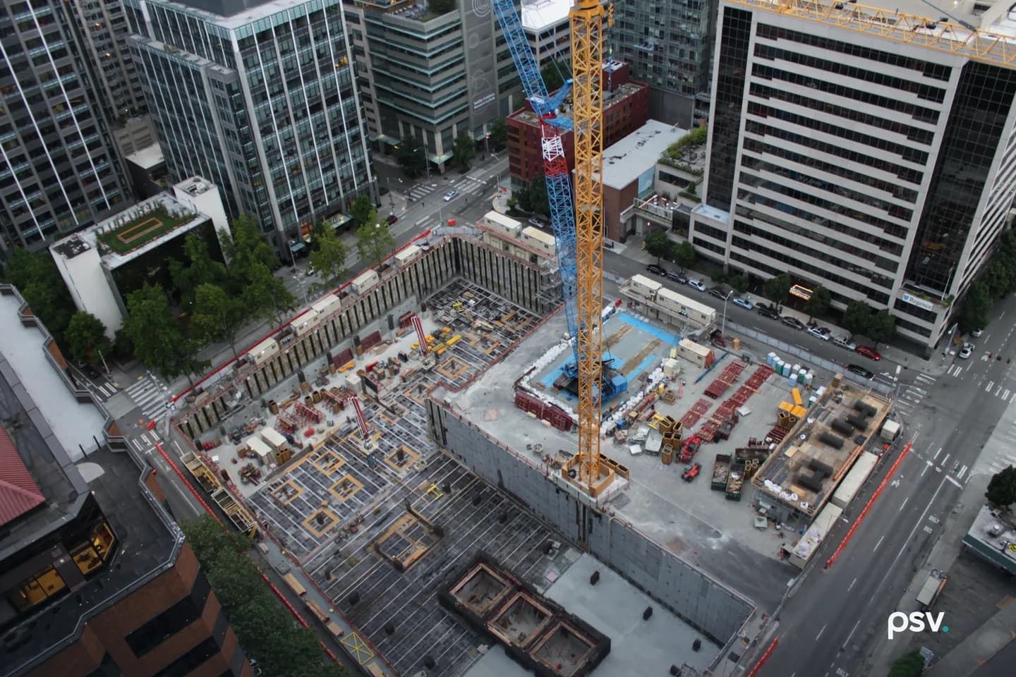 An overhead daylight view looking straight down into a downtown construction site on a city block: a yellow tower crane and a blue tower crane rise from the middle of the excavation, the left half of the pit is a grid of rebar and formwork for a foundation slab with scattered materials and small work trailers along a shored perimeter wall, the right half is a finished concrete deck crowded with stacked materials, red equipment and a blue-lined pad, and office and residential towers surround the block on every side with cars and crosswalks on the streets below. An overhead daylight view looking straight down into a downtown construction site on a city block: a yellow tower crane and a blue tower crane rise from the middle of the excavation, the left half of the pit is a grid of rebar and formwork for a foundation slab with scattered materials and small work trailers along a shored perimeter wall, the right half is a finished concrete deck crowded with stacked materials, red equipment and a blue-lined pad, and office and residential towers surround the block on every side with cars and crosswalks on the streets below.