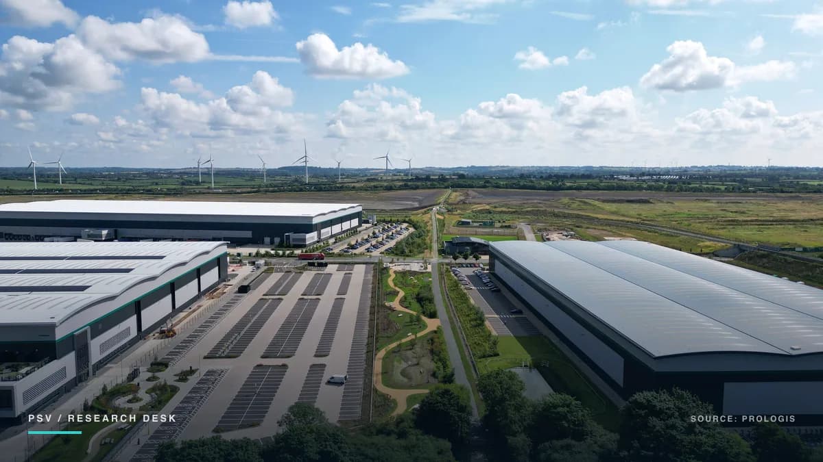 Aerial view of a UK logistics park: three long warehouse buildings with curved white roofs, two of them part-covered in solar panels, arranged around wide empty car parks and a landscaped drainage channel, with green farmland and a line of wind turbines on the horizon under a bright cloudy sky