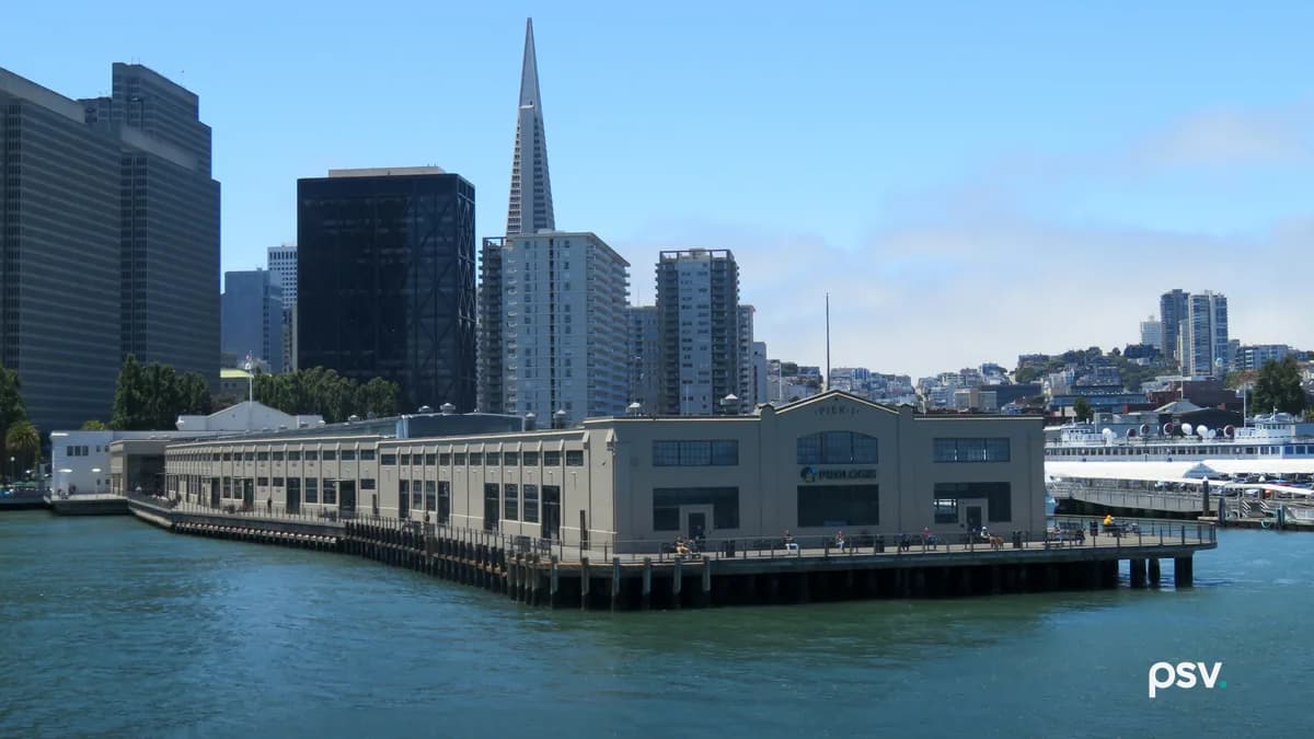 A long, pale two-storey pier shed on wooden pilings over green-blue bay water, with a PROLOGIS sign on its gabled end under the words PIER 1, people walking along the railed apron, the Transamerica Pyramid and other downtown San Francisco towers behind it, and a ferry docked at right under a clear blue sky.