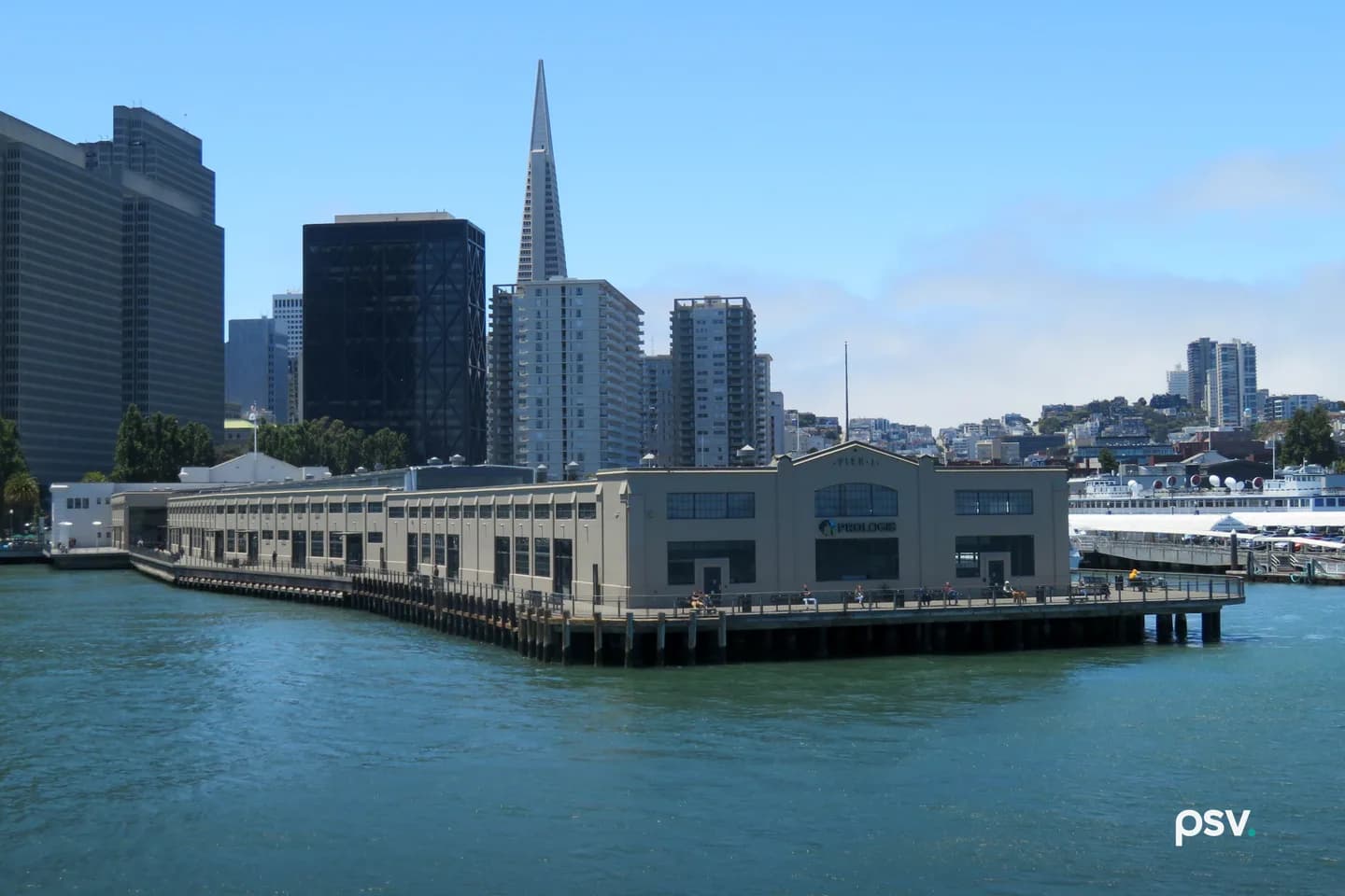 A long, pale two-storey pier shed on wooden pilings over green-blue bay water, with a PROLOGIS sign on its gabled end under the words PIER 1, people walking along the railed apron, the Transamerica Pyramid and other downtown San Francisco towers behind it, and a ferry docked at right under a clear blue sky. A long, pale two-storey pier shed on wooden pilings over green-blue bay water, with a PROLOGIS sign on its gabled end under the words PIER 1, people walking along the railed apron, the Transamerica Pyramid and other downtown San Francisco towers behind it, and a ferry docked at right under a clear blue sky.