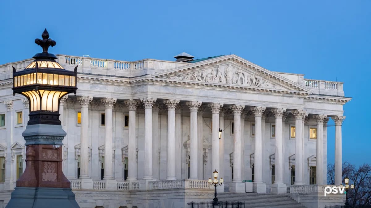 The east front of the Senate wing of the United States Capitol at dusk: a white marble portico of tall Corinthian columns under a sculpted pediment, wide steps rising from the plaza at right with lit lamp posts, a few lit windows, and an ornate lit lantern on a red granite and bronze post in the left foreground against a clear deep blue sky