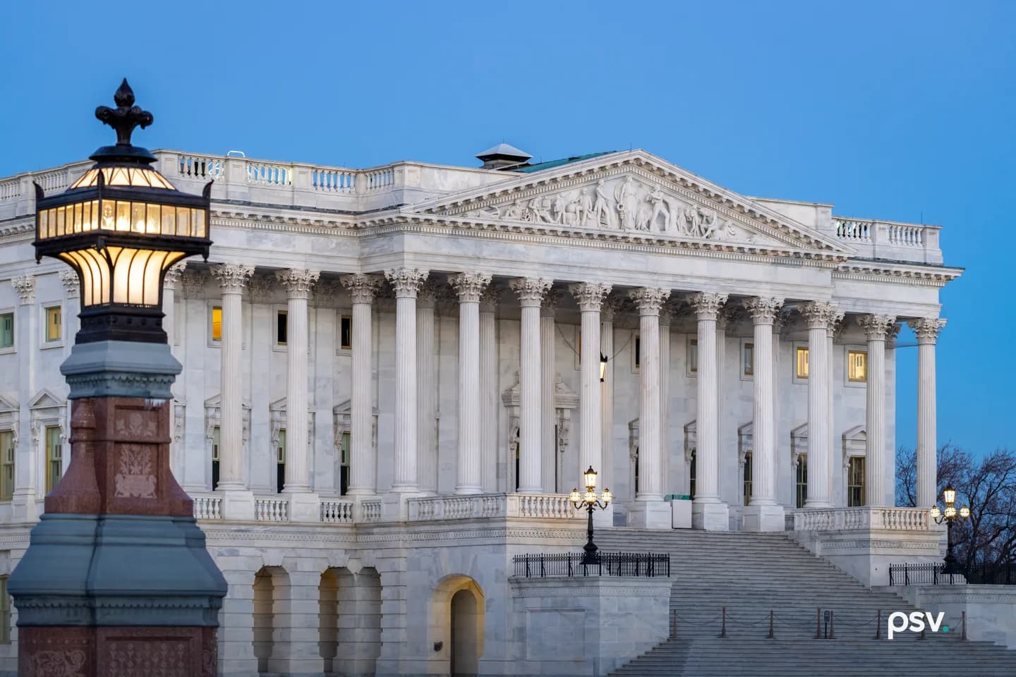 The east front of the Senate wing of the United States Capitol at dusk: a white marble portico of tall Corinthian columns under a sculpted pediment, wide steps rising from the plaza at right with lit lamp posts, a few lit windows, and an ornate lit lantern on a red granite and bronze post in the left foreground against a clear deep blue sky The east front of the Senate wing of the United States Capitol at dusk: a white marble portico of tall Corinthian columns under a sculpted pediment, wide steps rising from the plaza at right with lit lamp posts, a few lit windows, and an ornate lit lantern on a red granite and bronze post in the left foreground against a clear deep blue sky