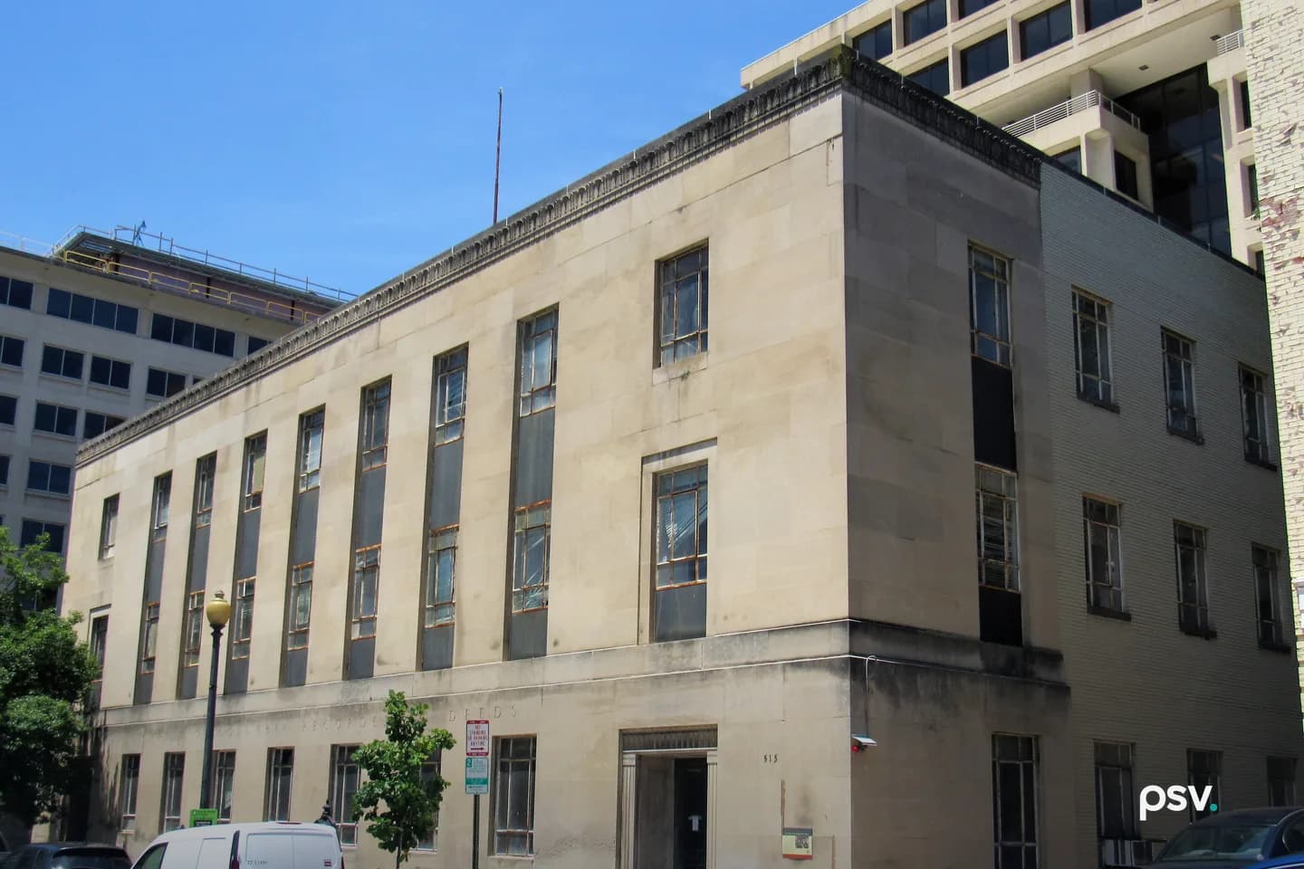 Street-level daylight view of the limestone District of Columbia Recorder of Deeds Building at 515 D Street NW in Washington, seen from the corner: three storeys of tall recessed windows in stone piers, a carved RECORDER OF DEEDS inscription running along the lintel course above the ground floor, a plain stone entrance surround at the corner, a darker brick wing on the right, and taller modern office buildings rising behind it under a clear blue sky Street-level daylight view of the limestone District of Columbia Recorder of Deeds Building at 515 D Street NW in Washington, seen from the corner: three storeys of tall recessed windows in stone piers, a carved RECORDER OF DEEDS inscription running along the lintel course above the ground floor, a plain stone entrance surround at the corner, a darker brick wing on the right, and taller modern office buildings rising behind it under a clear blue sky
