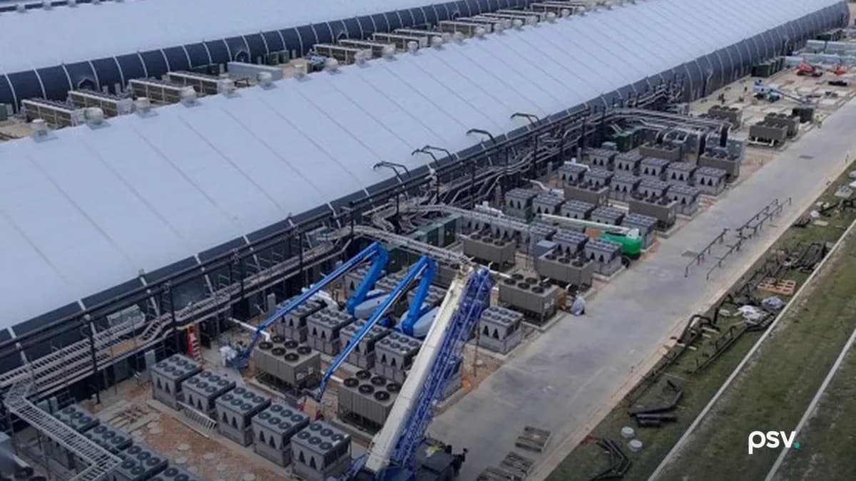 Aerial view of Riot’s Rockdale, Texas campus: two long white barrel-roofed data halls running to the horizon, ranks of grey air-cooled condenser units and transformer pads lining the service road beside them, a blue mobile crane and boom lifts at work mid-construction, coiled cable and pallets on the graded verge, with a power plant’s stacks visible in the distance
