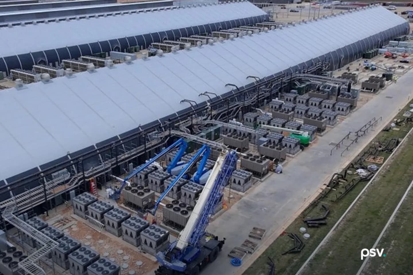 Aerial view of Riot’s Rockdale, Texas campus: two long white barrel-roofed data halls running to the horizon, ranks of grey air-cooled condenser units and transformer pads lining the service road beside them, a blue mobile crane and boom lifts at work mid-construction, coiled cable and pallets on the graded verge, with a power plant’s stacks visible in the distance Aerial view of Riot’s Rockdale, Texas campus: two long white barrel-roofed data halls running to the horizon, ranks of grey air-cooled condenser units and transformer pads lining the service road beside them, a blue mobile crane and boom lifts at work mid-construction, coiled cable and pallets on the graded verge, with a power plant’s stacks visible in the distance