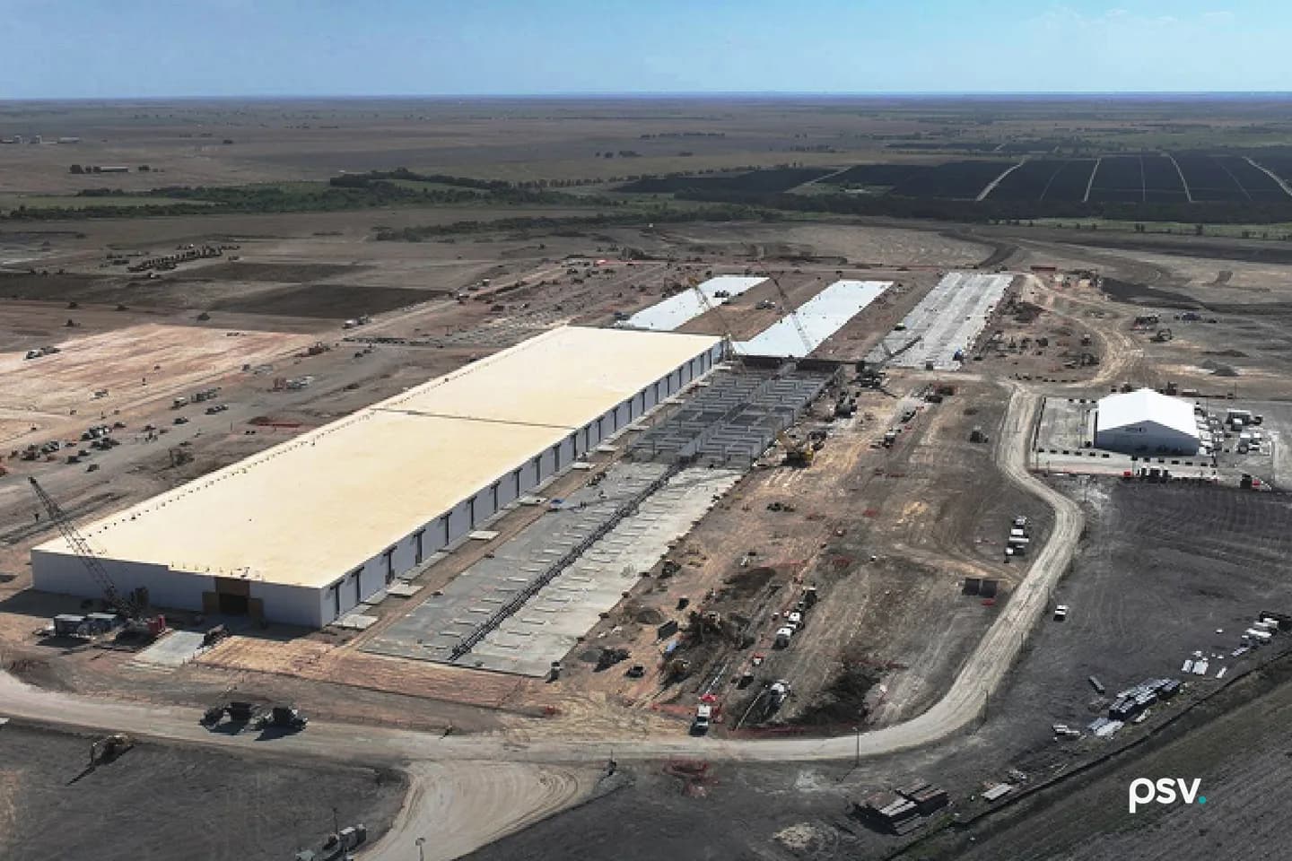 An aerial daylight photograph of a large data center campus under construction on flat rural land: a long single-storey building with a pale roof and white wall panels runs diagonally across the frame, beside it a row of exposed steel frames and concrete slabs marks buildings not yet enclosed, three more pale slabs extend toward the horizon with cranes working on them, a white tent structure and parked equipment sit to the right, a curving dirt haul road wraps the site, and dark rows of solar panels and open farmland stretch to the horizon under a blue sky. An aerial daylight photograph of a large data center campus under construction on flat rural land: a long single-storey building with a pale roof and white wall panels runs diagonally across the frame, beside it a row of exposed steel frames and concrete slabs marks buildings not yet enclosed, three more pale slabs extend toward the horizon with cranes working on them, a white tent structure and parked equipment sit to the right, a curving dirt haul road wraps the site, and dark rows of solar panels and open farmland stretch to the horizon under a blue sky.