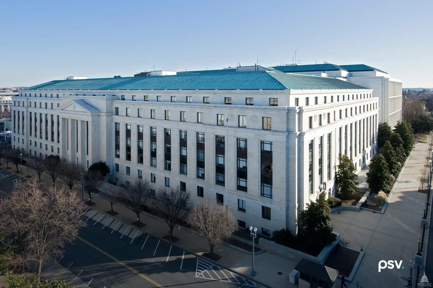 An elevated daylight view of the Dirksen Senate Office Building in Washington: a long, eight-story white marble building with a green copper hipped roof and a columned portico at its far end, bare winter trees and a parking lane along the street in front, evergreen shrubs and a wide plaza walkway at right, under a clear blue sky. An elevated daylight view of the Dirksen Senate Office Building in Washington: a long, eight-story white marble building with a green copper hipped roof and a columned portico at its far end, bare winter trees and a parking lane along the street in front, evergreen shrubs and a wide plaza walkway at right, under a clear blue sky.
