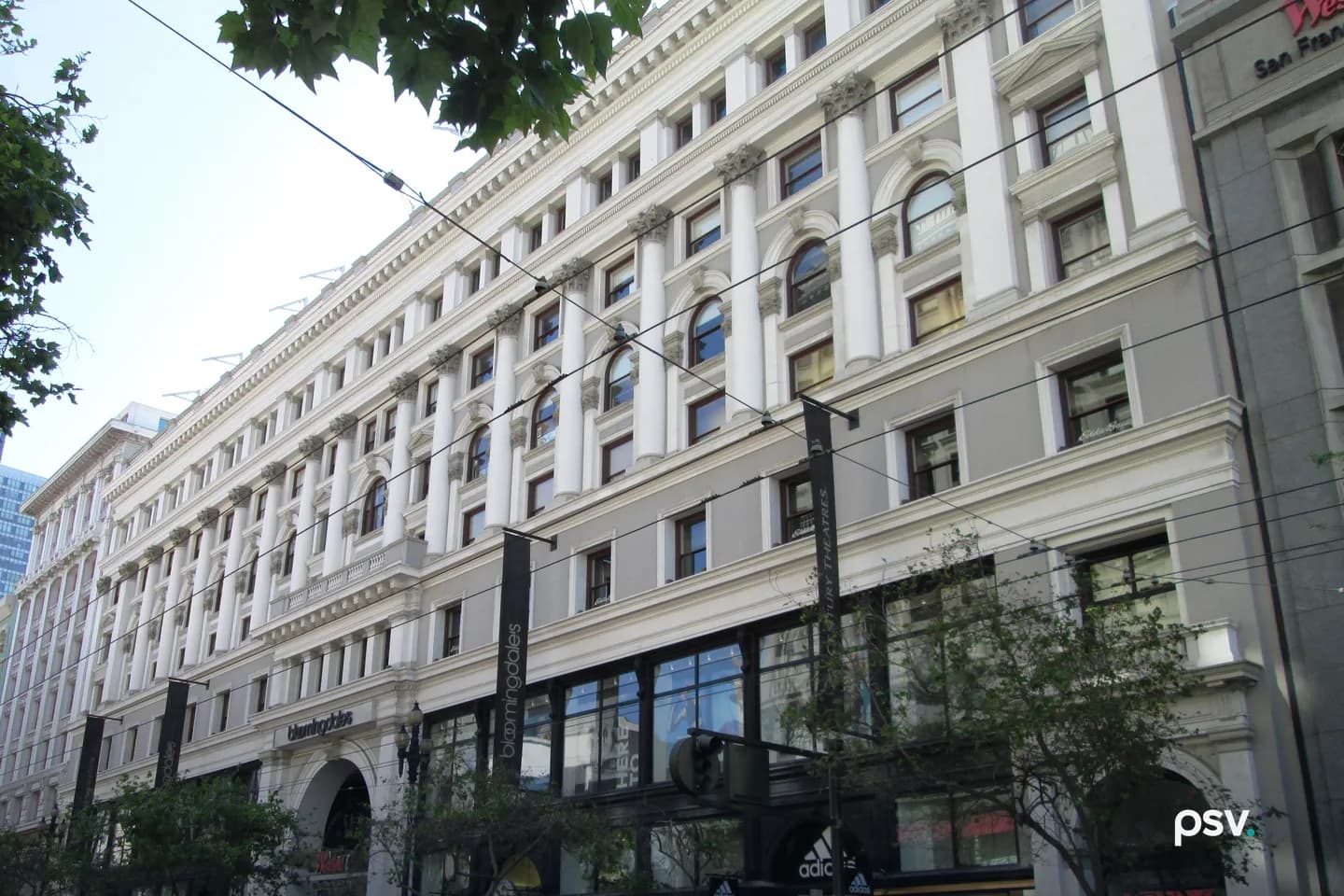 A white and gray seven-story classical building facade on Market Street in San Francisco with tall columns, arched windows and a cornice, a Bloomingdale’s sign over the ground-floor entrance, and overhead trolley wires crossing the frame. A white and gray seven-story classical building facade on Market Street in San Francisco with tall columns, arched windows and a cornice, a Bloomingdale’s sign over the ground-floor entrance, and overhead trolley wires crossing the frame.