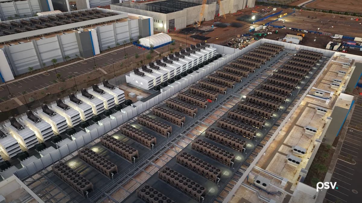 An aerial dusk view straight down onto the roof and yard of a data center campus: two long rows of rectangular heat rejection units, each packed with circular fans, racked on steel frames across a flat roof deck, with a parallel row of white generator enclosures topped by black exhaust stacks to the left, service walkways and pipe runs between them, an adjacent long white data hall at upper left, and a second hall still under construction at upper right with a yellow crawler crane, exposed steel framing, bare red earth and stacked materials.