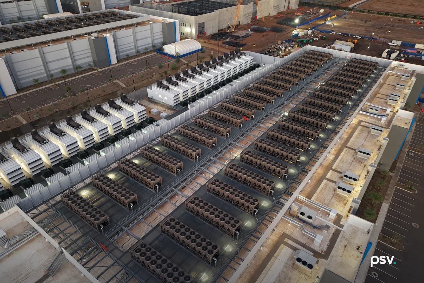 An aerial dusk view straight down onto the roof and yard of a data center campus: two long rows of rectangular heat rejection units, each packed with circular fans, racked on steel frames across a flat roof deck, with a parallel row of white generator enclosures topped by black exhaust stacks to the left, service walkways and pipe runs between them, an adjacent long white data hall at upper left, and a second hall still under construction at upper right with a yellow crawler crane, exposed steel framing, bare red earth and stacked materials. An aerial dusk view straight down onto the roof and yard of a data center campus: two long rows of rectangular heat rejection units, each packed with circular fans, racked on steel frames across a flat roof deck, with a parallel row of white generator enclosures topped by black exhaust stacks to the left, service walkways and pipe runs between them, an adjacent long white data hall at upper left, and a second hall still under construction at upper right with a yellow crawler crane, exposed steel framing, bare red earth and stacked materials.
