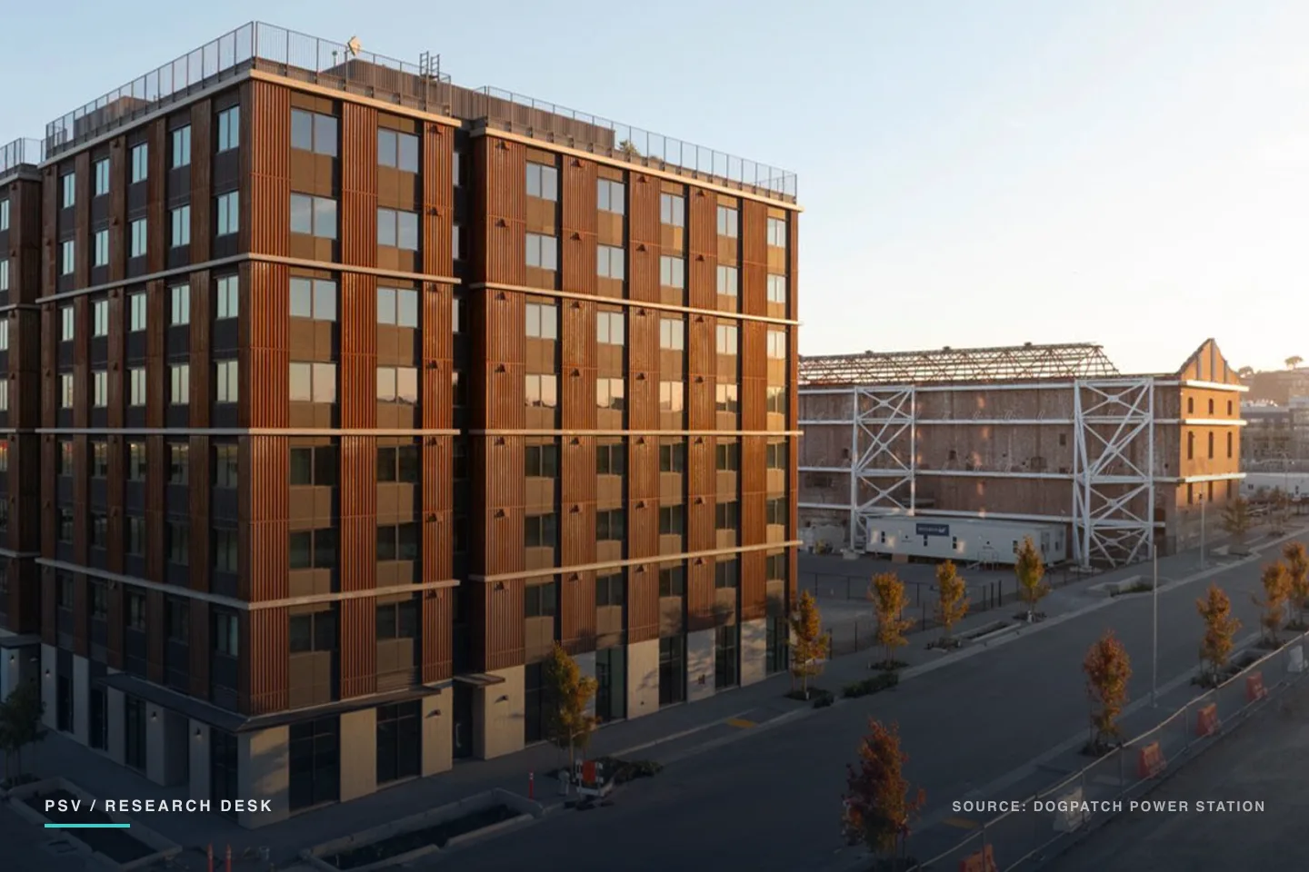 An eight-story apartment building clad in ribbed corten steel stands at a street corner in late afternoon light, with young street trees along the sidewalk and the white steel bracing of the historic Station A power plant shell visible behind it An eight-story apartment building clad in ribbed corten steel stands at a street corner in late afternoon light, with young street trees along the sidewalk and the white steel bracing of the historic Station A power plant shell visible behind it