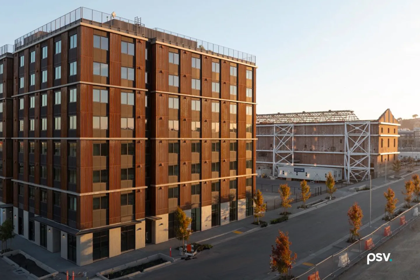 An eight-story apartment building clad in ribbed corten steel stands at a street corner in late afternoon light, with young street trees along the sidewalk and the white steel bracing of the historic Station A power plant shell visible behind it An eight-story apartment building clad in ribbed corten steel stands at a street corner in late afternoon light, with young street trees along the sidewalk and the white steel bracing of the historic Station A power plant shell visible behind it