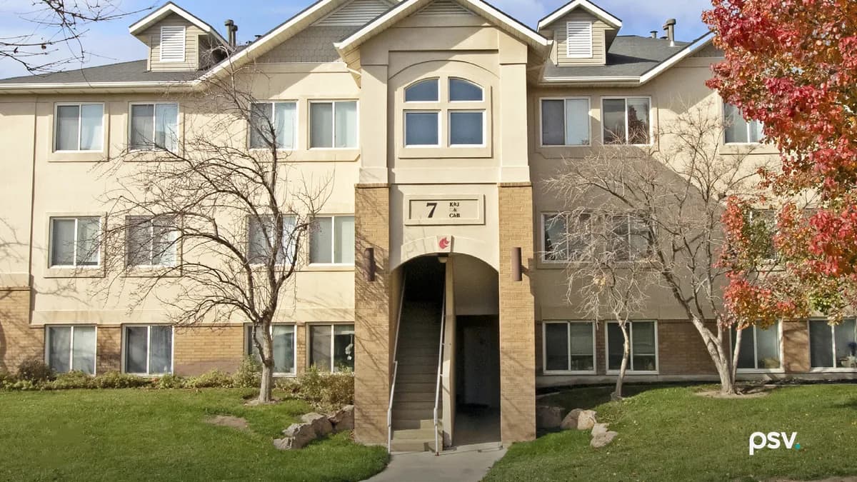 A three-story garden apartment building with cream stucco and tan brick, a gabled entry bay with an arched window above a covered stairwell marked with the building number 7, bare trees and one tree in full red autumn colour on a mown lawn in front
