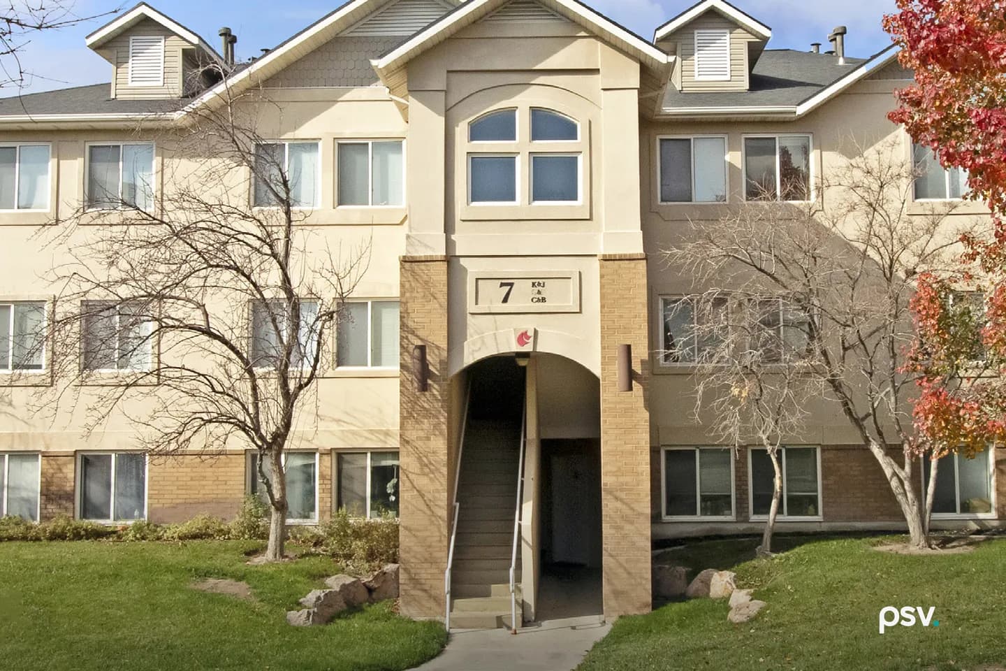 A three-story garden apartment building with cream stucco and tan brick, a gabled entry bay with an arched window above a covered stairwell marked with the building number 7, bare trees and one tree in full red autumn colour on a mown lawn in front A three-story garden apartment building with cream stucco and tan brick, a gabled entry bay with an arched window above a covered stairwell marked with the building number 7, bare trees and one tree in full red autumn colour on a mown lawn in front