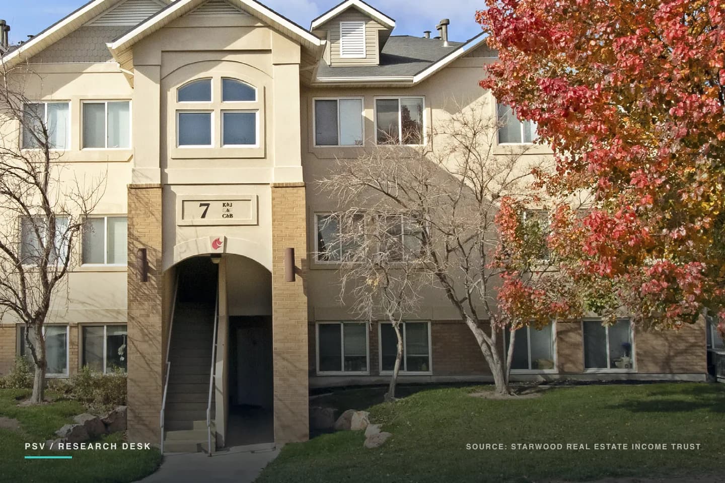 A three-story garden apartment building with cream stucco and tan brick, a gabled entry bay with an arched window above a covered stairwell marked with the building number 7, bare trees and one tree in full red autumn colour on a mown lawn in front A three-story garden apartment building with cream stucco and tan brick, a gabled entry bay with an arched window above a covered stairwell marked with the building number 7, bare trees and one tree in full red autumn colour on a mown lawn in front