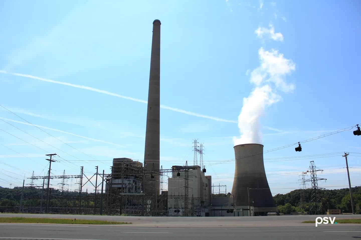 A power plant seen from across a road under a blue sky, with a tall concrete smokestack at the center, a hyperbolic cooling tower releasing white vapor at the right, boiler buildings between them, and a switchyard of steel structures and transmission towers along the front. A power plant seen from across a road under a blue sky, with a tall concrete smokestack at the center, a hyperbolic cooling tower releasing white vapor at the right, boiler buildings between them, and a switchyard of steel structures and transmission towers along the front.