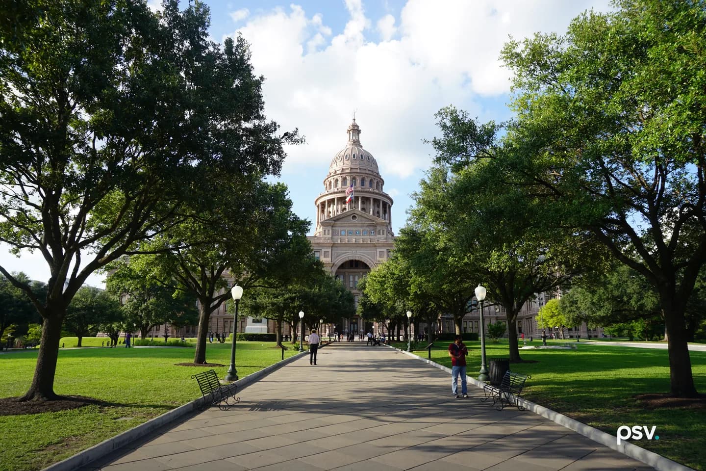 A daylight photograph of the Texas State Capitol in Austin, a pink granite building with a tall columned dome flying the United States and Texas flags, seen at the end of a long paved walkway lined with iron lamp posts and green benches, framed by large live oak trees and wide green lawns, with a few pedestrians small in the distance under a blue sky with white clouds. A daylight photograph of the Texas State Capitol in Austin, a pink granite building with a tall columned dome flying the United States and Texas flags, seen at the end of a long paved walkway lined with iron lamp posts and green benches, framed by large live oak trees and wide green lawns, with a few pedestrians small in the distance under a blue sky with white clouds.