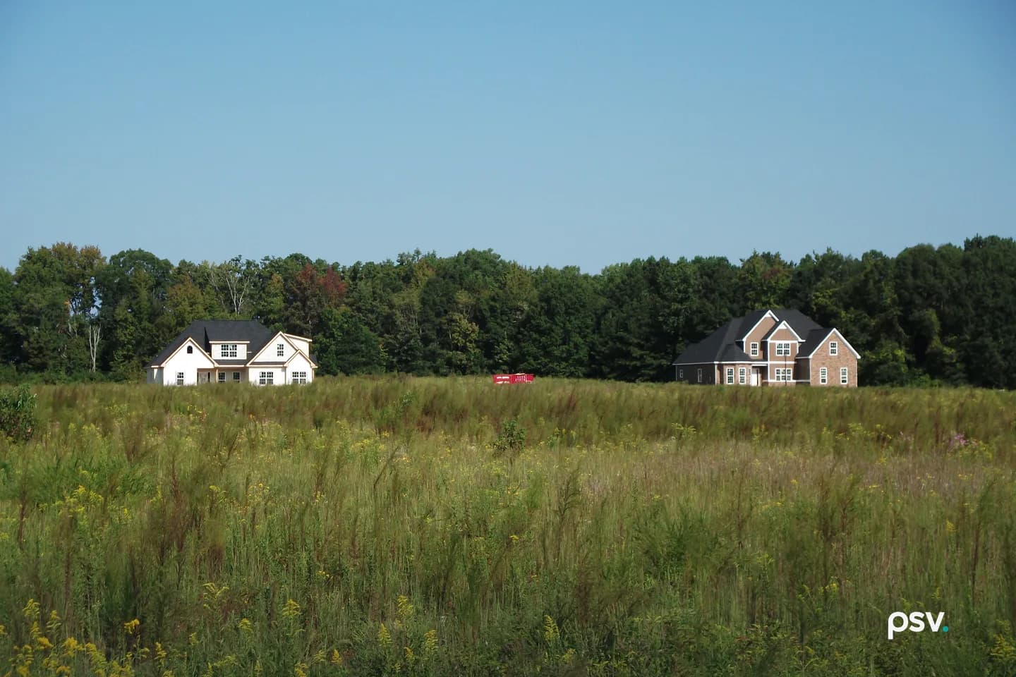 Two newly built two-story houses, one white with a dark roof and one brick and gray, standing apart at the far edge of a field of tall grass and yellow wildflowers in front of a tree line under a clear blue sky, with a red dumpster between them. Two newly built two-story houses, one white with a dark roof and one brick and gray, standing apart at the far edge of a field of tall grass and yellow wildflowers in front of a tree line under a clear blue sky, with a red dumpster between them.