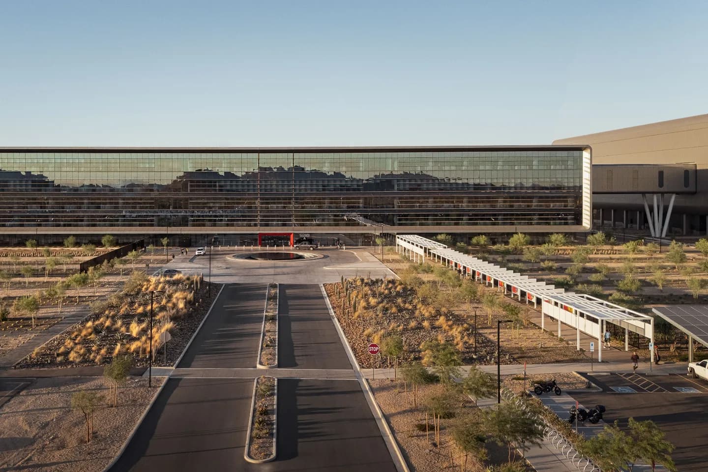 TSMC's semiconductor campus in north Phoenix: the long mirrored-glass administration building fronting a landscaped desert entry drive and roundabout, with fabrication buildings beyond, at golden hour TSMC's semiconductor campus in north Phoenix: the long mirrored-glass administration building fronting a landscaped desert entry drive and roundabout, with fabrication buildings beyond, at golden hour
