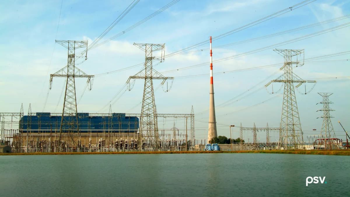 Daylight view across a still cooling pond toward a Tennessee Valley Authority generating plant: four lattice steel transmission towers carrying multiple high voltage circuits, a crowded switchyard of insulators and breakers along the far bank, a long blue-clad turbine building at left, and a tall red and white banded stack at center under a pale blue sky