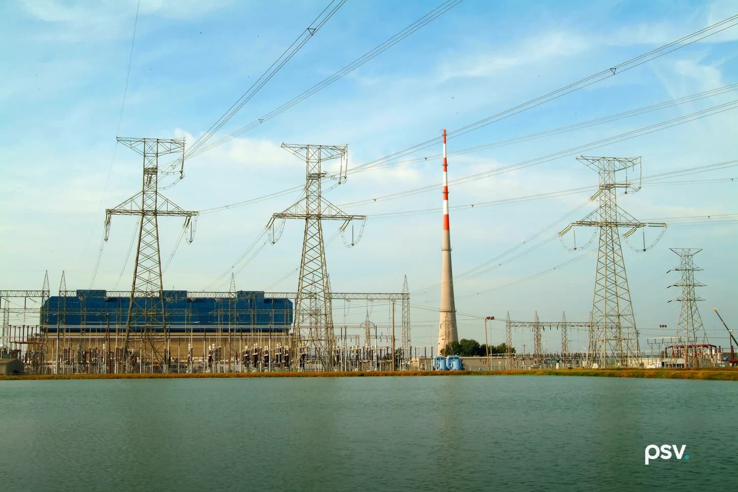 Daylight view across a still cooling pond toward a Tennessee Valley Authority generating plant: four lattice steel transmission towers carrying multiple high voltage circuits, a crowded switchyard of insulators and breakers along the far bank, a long blue-clad turbine building at left, and a tall red and white banded stack at center under a pale blue sky Daylight view across a still cooling pond toward a Tennessee Valley Authority generating plant: four lattice steel transmission towers carrying multiple high voltage circuits, a crowded switchyard of insulators and breakers along the far bank, a long blue-clad turbine building at left, and a tall red and white banded stack at center under a pale blue sky