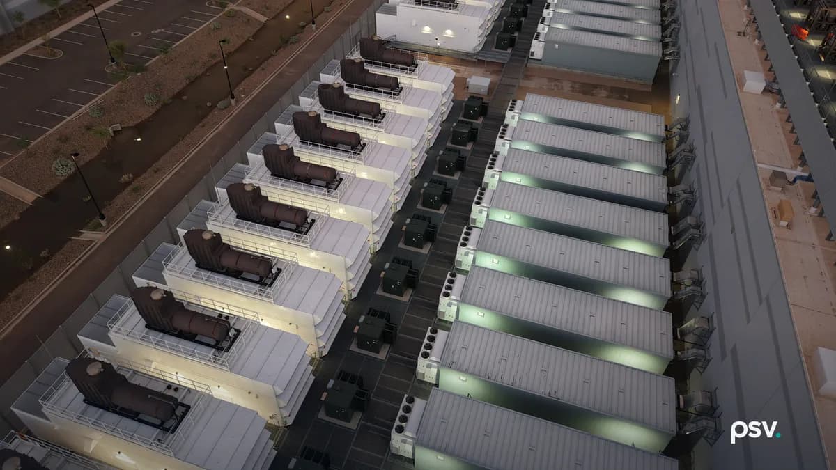 An aerial dusk view down onto the service yard of a data center campus in the desert: a long diagonal row of white generator enclosures, each topped by a dark exhaust silencer on a railed steel platform, running beside a parallel row of long ribbed equipment houses lit from within, with dark switchgear and transformer cabinets lined up in the paved lane between the two rows, a tall perimeter wall, an empty striped parking lot and gravel desert landscaping at left, and the flank of a data hall with a louvered wall at right.
