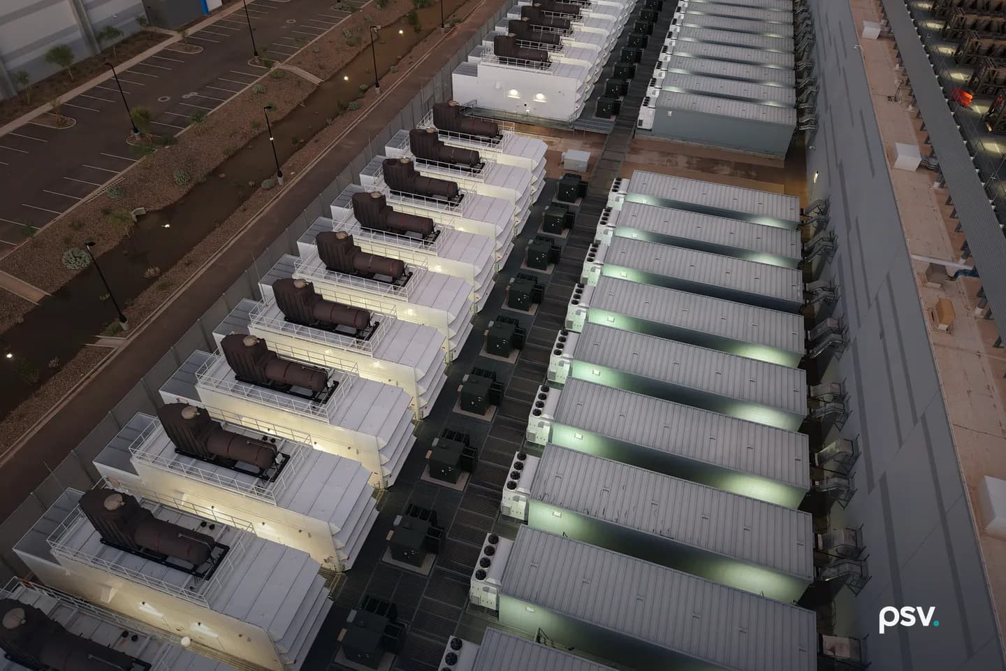 An aerial dusk view down onto the service yard of a data center campus in the desert: a long diagonal row of white generator enclosures, each topped by a dark exhaust silencer on a railed steel platform, running beside a parallel row of long ribbed equipment houses lit from within, with dark switchgear and transformer cabinets lined up in the paved lane between the two rows, a tall perimeter wall, an empty striped parking lot and gravel desert landscaping at left, and the flank of a data hall with a louvered wall at right. An aerial dusk view down onto the service yard of a data center campus in the desert: a long diagonal row of white generator enclosures, each topped by a dark exhaust silencer on a railed steel platform, running beside a parallel row of long ribbed equipment houses lit from within, with dark switchgear and transformer cabinets lined up in the paved lane between the two rows, a tall perimeter wall, an empty striped parking lot and gravel desert landscaping at left, and the flank of a data hall with a louvered wall at right.