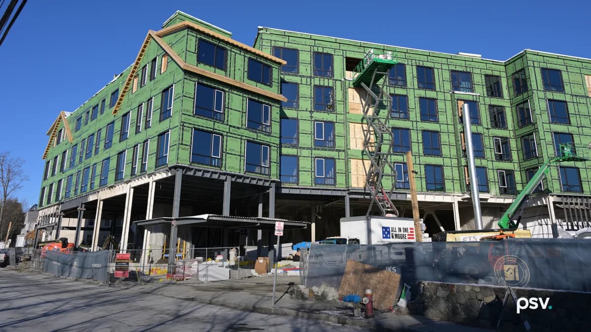 A five-storey apartment building under construction beneath a clear blue sky, its upper four floors wrapped in bright green sheathing with dark windows installed, standing on exposed steel columns over an open ground level, with a scissor lift raised to the top floor at the center, a green boom lift at the right, a white box truck parked in front, and a fenced site with stacked materials, a red fire hydrant and a low stone wall along the street.