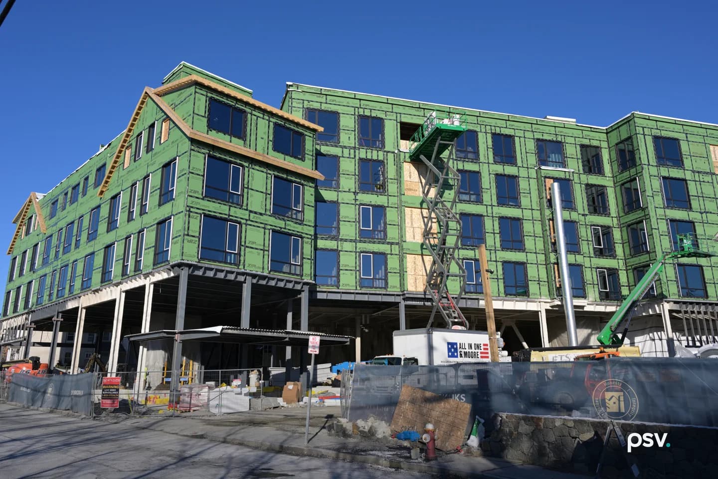 A five-storey apartment building under construction beneath a clear blue sky, its upper four floors wrapped in bright green sheathing with dark windows installed, standing on exposed steel columns over an open ground level, with a scissor lift raised to the top floor at the center, a green boom lift at the right, a white box truck parked in front, and a fenced site with stacked materials, a red fire hydrant and a low stone wall along the street. A five-storey apartment building under construction beneath a clear blue sky, its upper four floors wrapped in bright green sheathing with dark windows installed, standing on exposed steel columns over an open ground level, with a scissor lift raised to the top floor at the center, a green boom lift at the right, a white box truck parked in front, and a fenced site with stacked materials, a red fire hydrant and a low stone wall along the street.