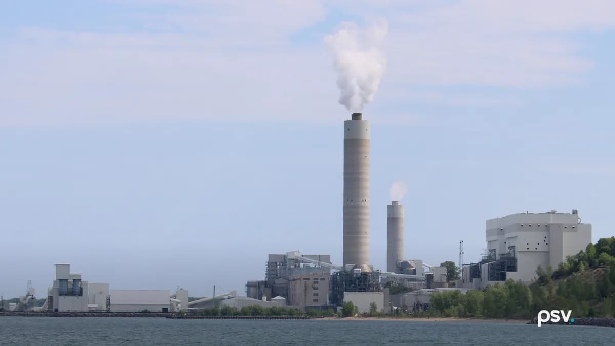A daylight view across Lake Michigan toward a large lakeside power station: one tall pale concrete chimney venting a white plume at center with a shorter second stack beside it, a low grey and beige complex of boiler houses, conveyor galleries and steel pipe racks spread along the shoreline below, a long white materials handling building and a dock wall at left, a bluff of green trees rising at right, and a strip of sandy beach and grey-green water across the foreground under a hazy blue sky.