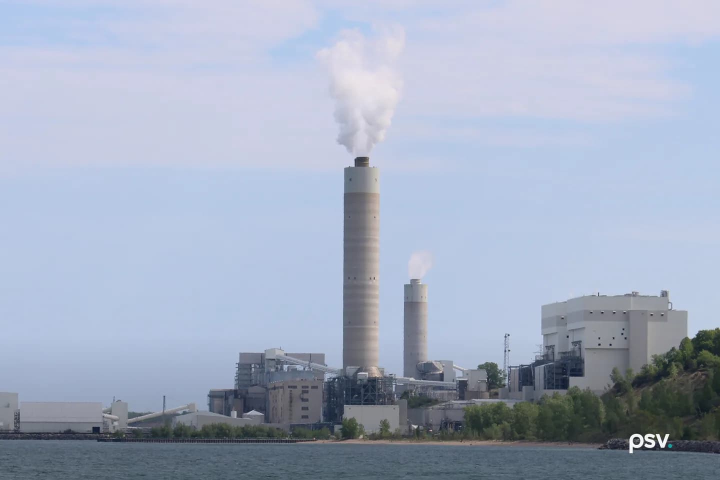 A daylight view across Lake Michigan toward a large lakeside power station: one tall pale concrete chimney venting a white plume at center with a shorter second stack beside it, a low grey and beige complex of boiler houses, conveyor galleries and steel pipe racks spread along the shoreline below, a long white materials handling building and a dock wall at left, a bluff of green trees rising at right, and a strip of sandy beach and grey-green water across the foreground under a hazy blue sky. A daylight view across Lake Michigan toward a large lakeside power station: one tall pale concrete chimney venting a white plume at center with a shorter second stack beside it, a low grey and beige complex of boiler houses, conveyor galleries and steel pipe racks spread along the shoreline below, a long white materials handling building and a dock wall at left, a bluff of green trees rising at right, and a strip of sandy beach and grey-green water across the foreground under a hazy blue sky.