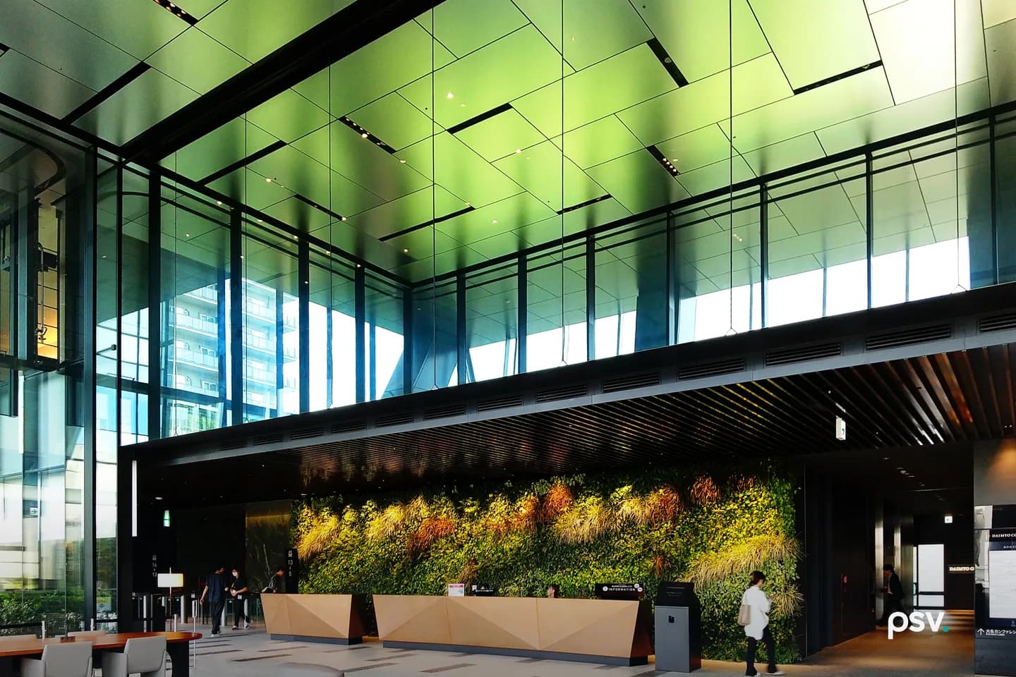 The daylit ground-floor lobby of a modern office tower, with a full-height glass curtain wall, a green-toned metal panel ceiling, a long planted living wall behind a folded-timber reception desk, and a few people crossing the floor at a distance. The daylit ground-floor lobby of a modern office tower, with a full-height glass curtain wall, a green-toned metal panel ceiling, a long planted living wall behind a folded-timber reception desk, and a few people crossing the floor at a distance.