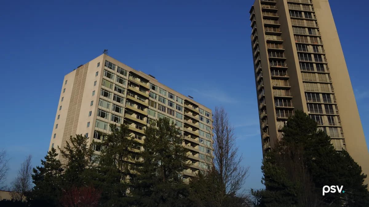Two residential high-rise towers with rows of balconies and windows, a lower slab-shaped tower on the left and a taller narrow tower on the right, seen from below against a clear blue sky with evergreen trees in the foreground.