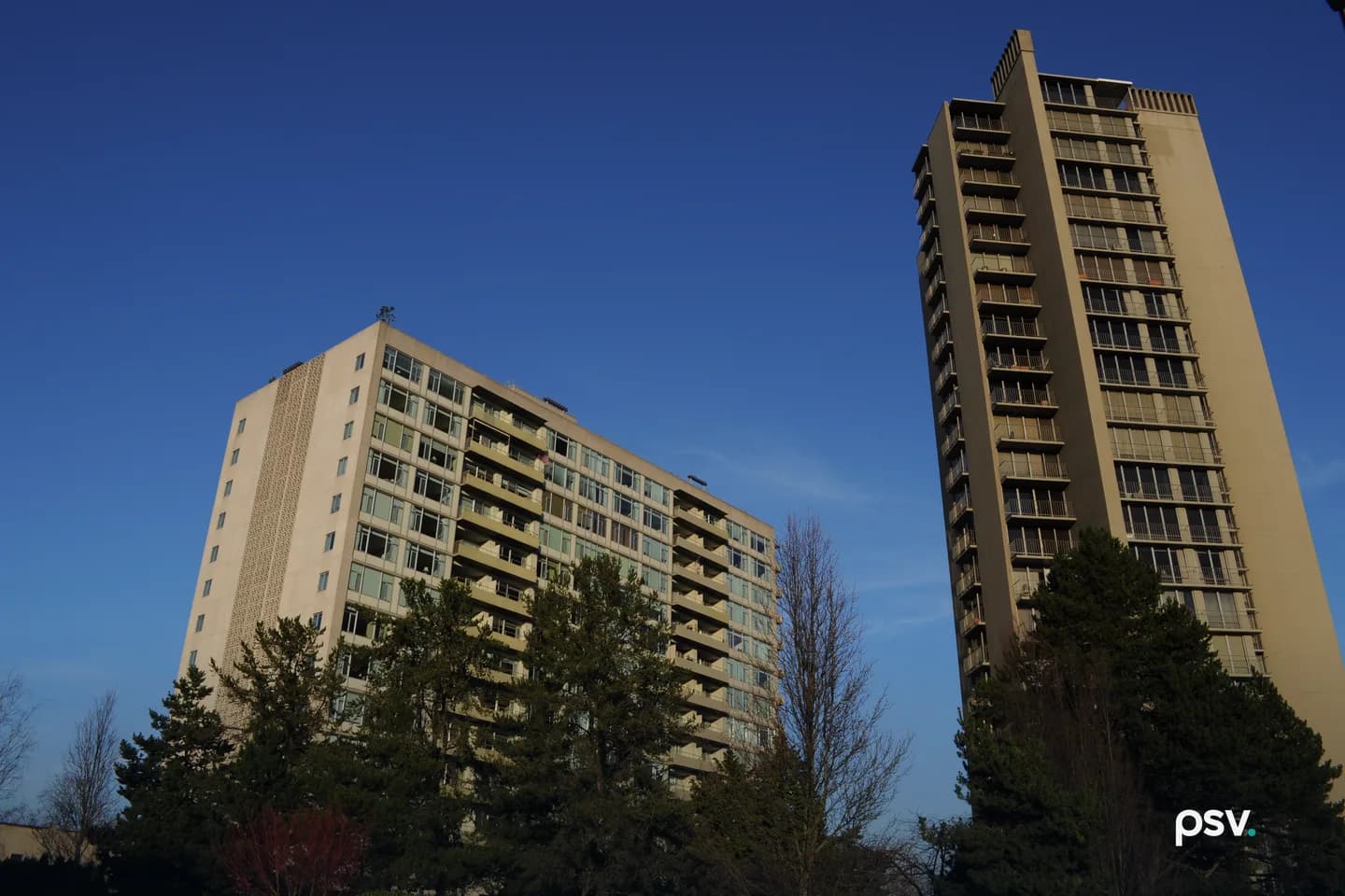 Two residential high-rise towers with rows of balconies and windows, a lower slab-shaped tower on the left and a taller narrow tower on the right, seen from below against a clear blue sky with evergreen trees in the foreground. Two residential high-rise towers with rows of balconies and windows, a lower slab-shaped tower on the left and a taller narrow tower on the right, seen from below against a clear blue sky with evergreen trees in the foreground.