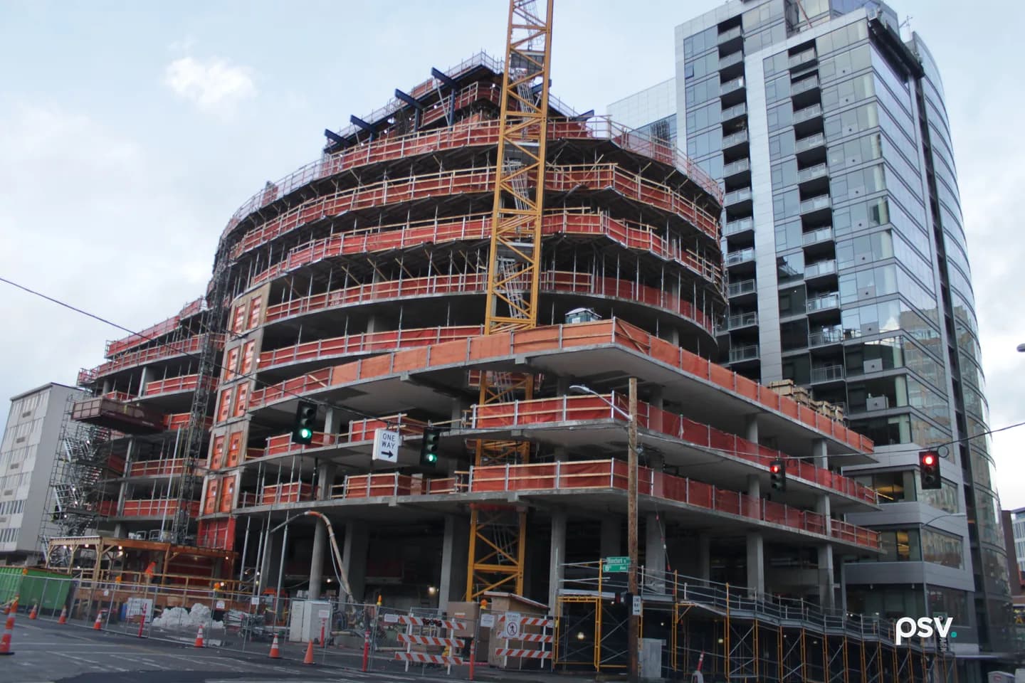 A curved apartment tower under construction on a Seattle street corner, with concrete floors wrapped in orange safety netting, a yellow tower crane, and a finished glass residential tower with balconies standing beside it. A curved apartment tower under construction on a Seattle street corner, with concrete floors wrapped in orange safety netting, a yellow tower crane, and a finished glass residential tower with balconies standing beside it.