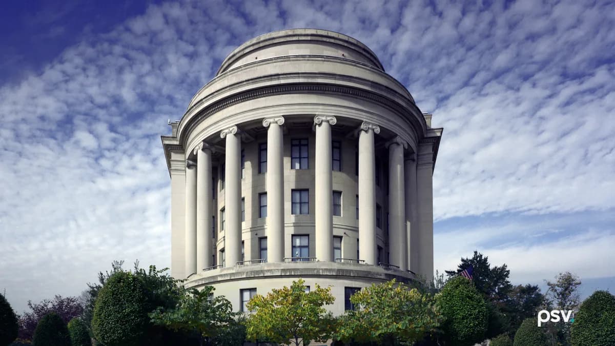 A daylight front view of the curved limestone west facade of the Federal Trade Commission Building in Washington, DC: a semicircular colonnade of tall Ionic columns beneath a stepped drum and shallow dome, with clipped trees and a low hedge across the foreground and a mackerel sky above.