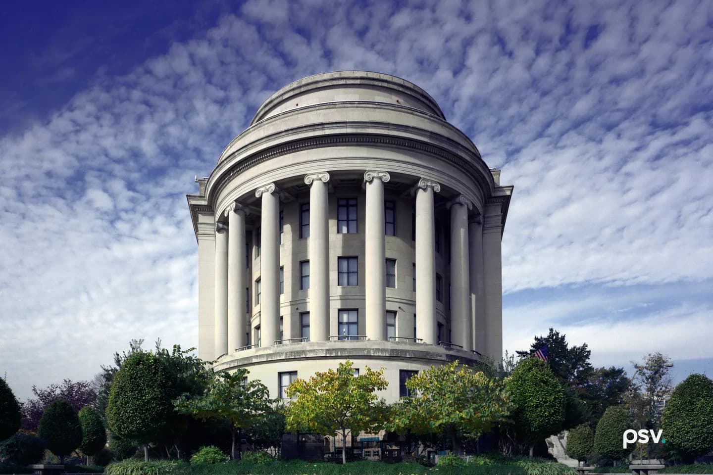 A daylight front view of the curved limestone west facade of the Federal Trade Commission Building in Washington, DC: a semicircular colonnade of tall Ionic columns beneath a stepped drum and shallow dome, with clipped trees and a low hedge across the foreground and a mackerel sky above. A daylight front view of the curved limestone west facade of the Federal Trade Commission Building in Washington, DC: a semicircular colonnade of tall Ionic columns beneath a stepped drum and shallow dome, with clipped trees and a low hedge across the foreground and a mackerel sky above.